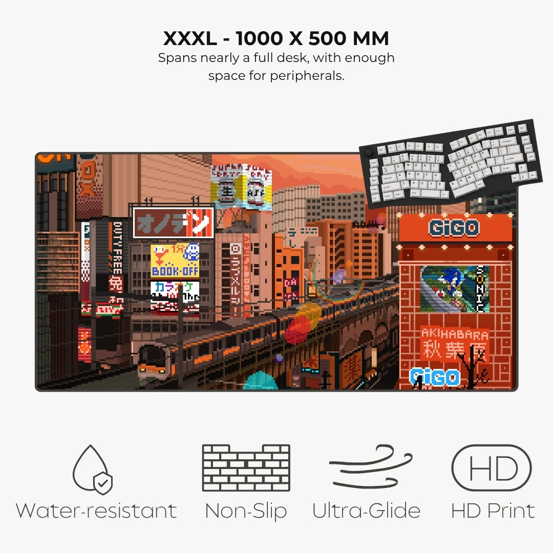 A large desk mat featuring a colorful pixel-art city scene with Japanese signage and a train, shown on a light gray background with text reading 'XXXL - 1000 X 500 MM' and feature icons for water resistance, non-slip, ultra-glide, and HD print.