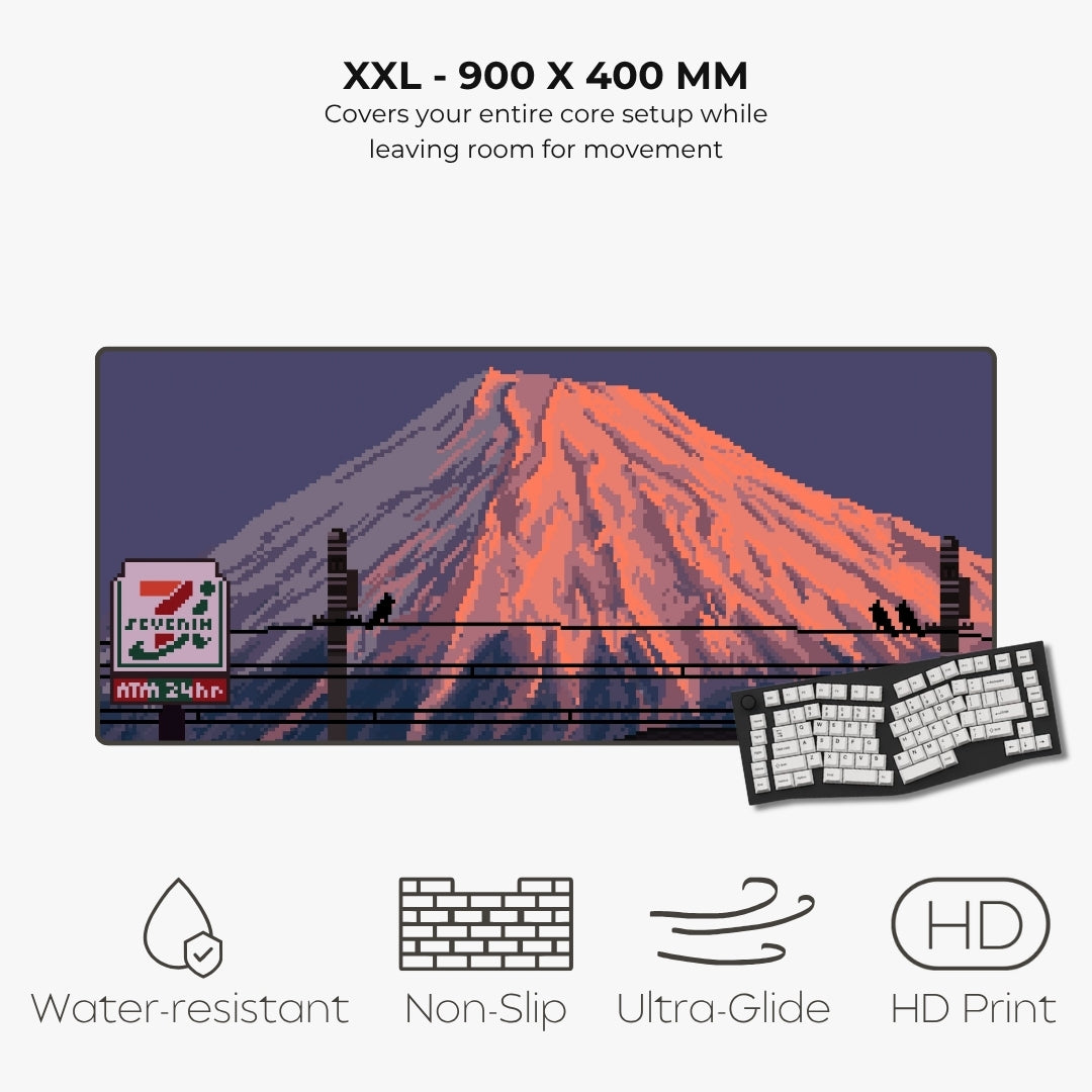 A large rectangular desk mat or mouse pad with a pixel-art style mount Fuji scene, shown on a light gray background with text reading 'XXL - 900 X 400 MM' and feature icons for water resistance, non-slip, ultra-glide, and HD print. A keyboard and mouse are partially visible underneath as scale/demo props.