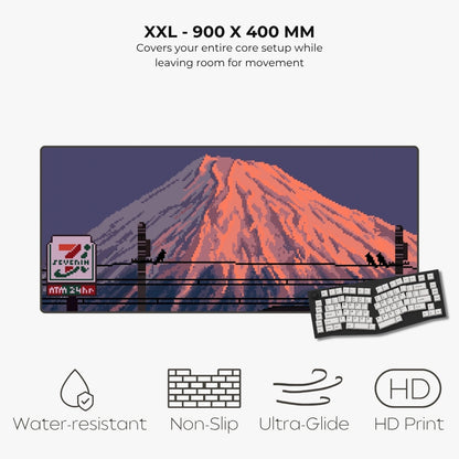 A large rectangular desk mat or mouse pad with a pixel-art style mount Fuji scene, shown on a light gray background with text reading 'XXL - 900 X 400 MM' and feature icons for water resistance, non-slip, ultra-glide, and HD print. A keyboard and mouse are partially visible underneath as scale/demo props.
