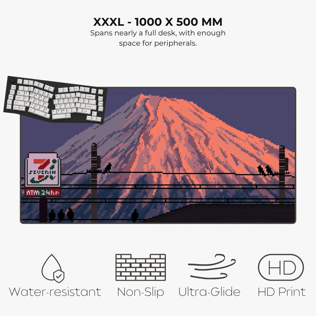 A large XXXL desk mat featuring a pixel-art style illustration of Mount Fuji with a 7-Eleven sign in the foreground, shown on a light gray background with text describing its size and features.