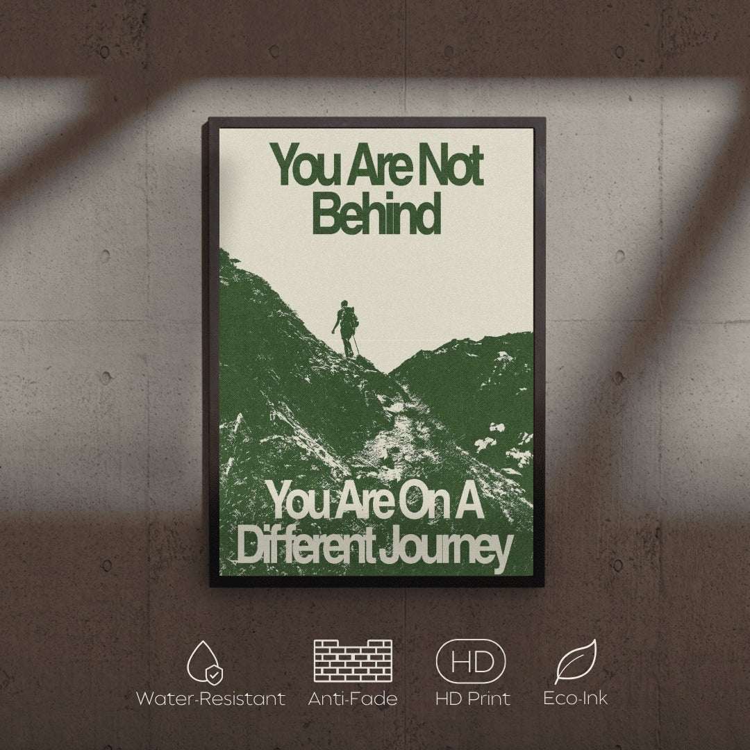Framed motivational poster with a silhouette of a person on a mountain and text 'You Are Not Behind, You Are On A Different Journey' on a textured wall.