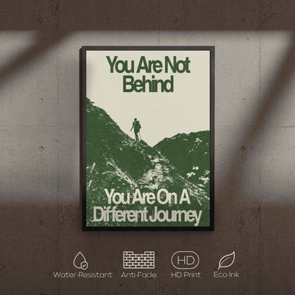 Framed motivational poster with a silhouette of a person on a mountain and text 'You Are Not Behind, You Are On A Different Journey' on a textured wall.