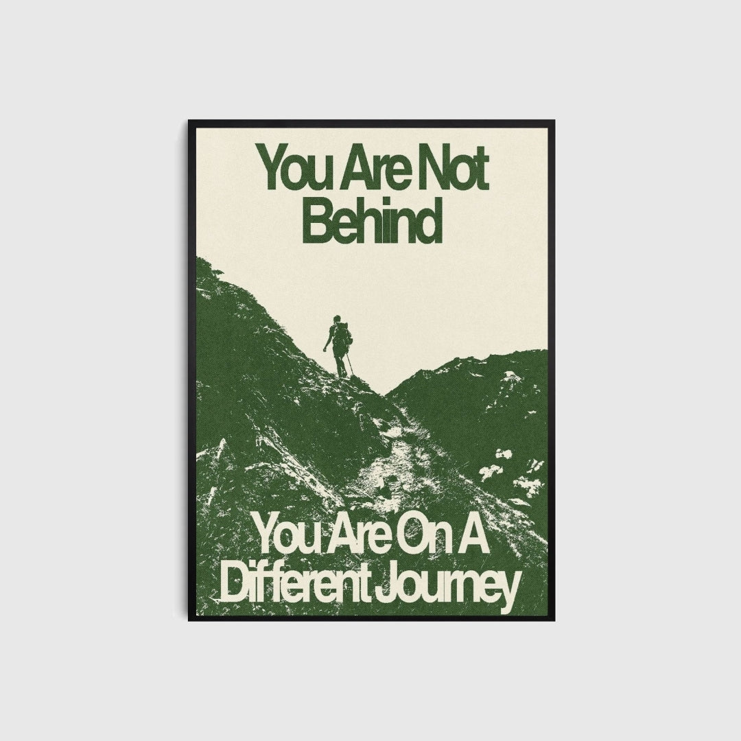 Framed motivational poster with a silhouette of a person on a mountain and text 'You Are Not Behind, You Are On A Different Journey' on a blank wall.