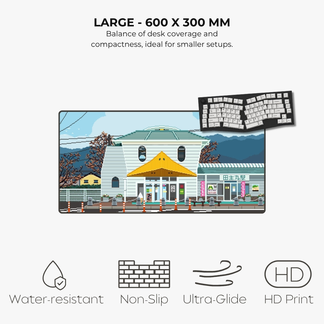 A large desk mat featuring a colorful pixel-art illustration of a Japanese street scene with a white building, yellow awning, blue sky, and mountains. The image includes marketing text reading 'LARGE - 600 X 300 MM' and feature icons for water-resistant, non-slip, ultra-glide, and HD print.