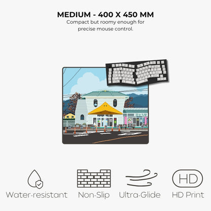 An illustrated pixel art mouse pad shown on a light gray background with marketing text above and feature icons below. The pad features a colorful cartoon street scene with buildings, trees, and a yellow awning.