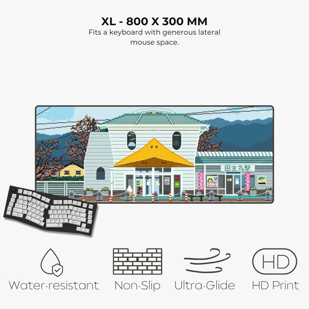 An XL-sized keyboard mouse pad featuring a colorful pixel-art illustration of a building and street scene. The pad is shown with a keyboard partially visible underneath to demonstrate fit, on a light gray background with text reading 'XL - 800 X 300 MM' and feature icons for water resistance, non-slip, ultra glide, and HD print.