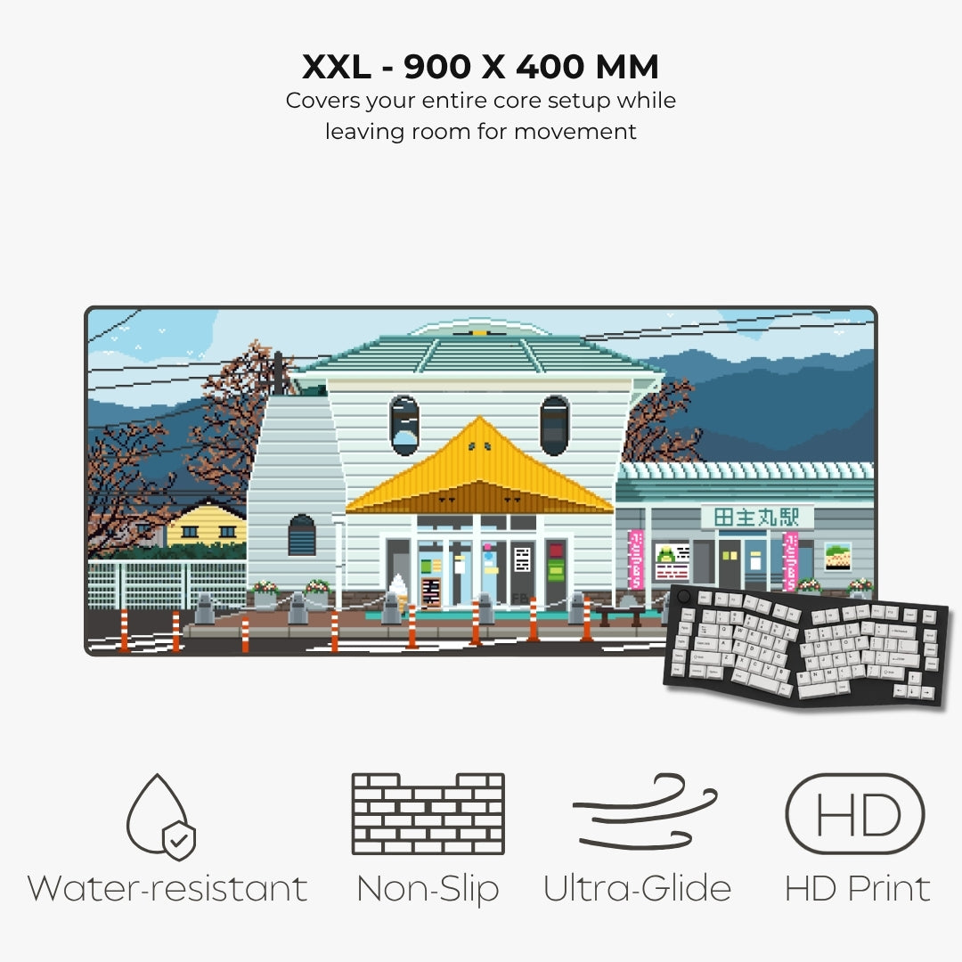 An XXL-sized printed desk mat featuring a pixel-art illustration of a building and storefront, shown on a light gray background with text highlighting its 900 x 400 mm size and features like water resistance, non-slip backing, ultra-glide edge, and HD print. A keyboard is pictured on the right as a usage example.